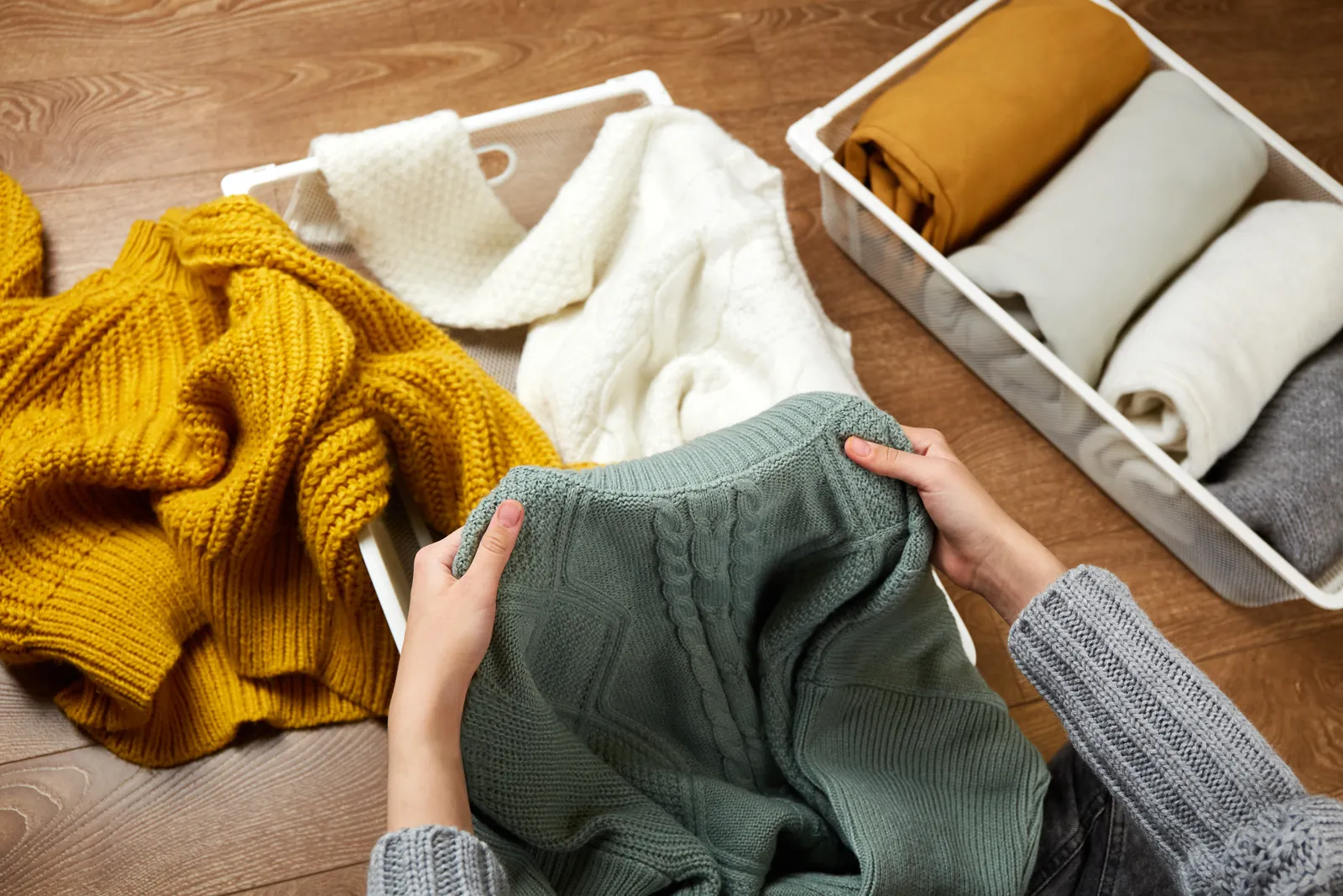 Hands folding a green knit sweater into a storage basket beside neat stacks of fall layers on the floor