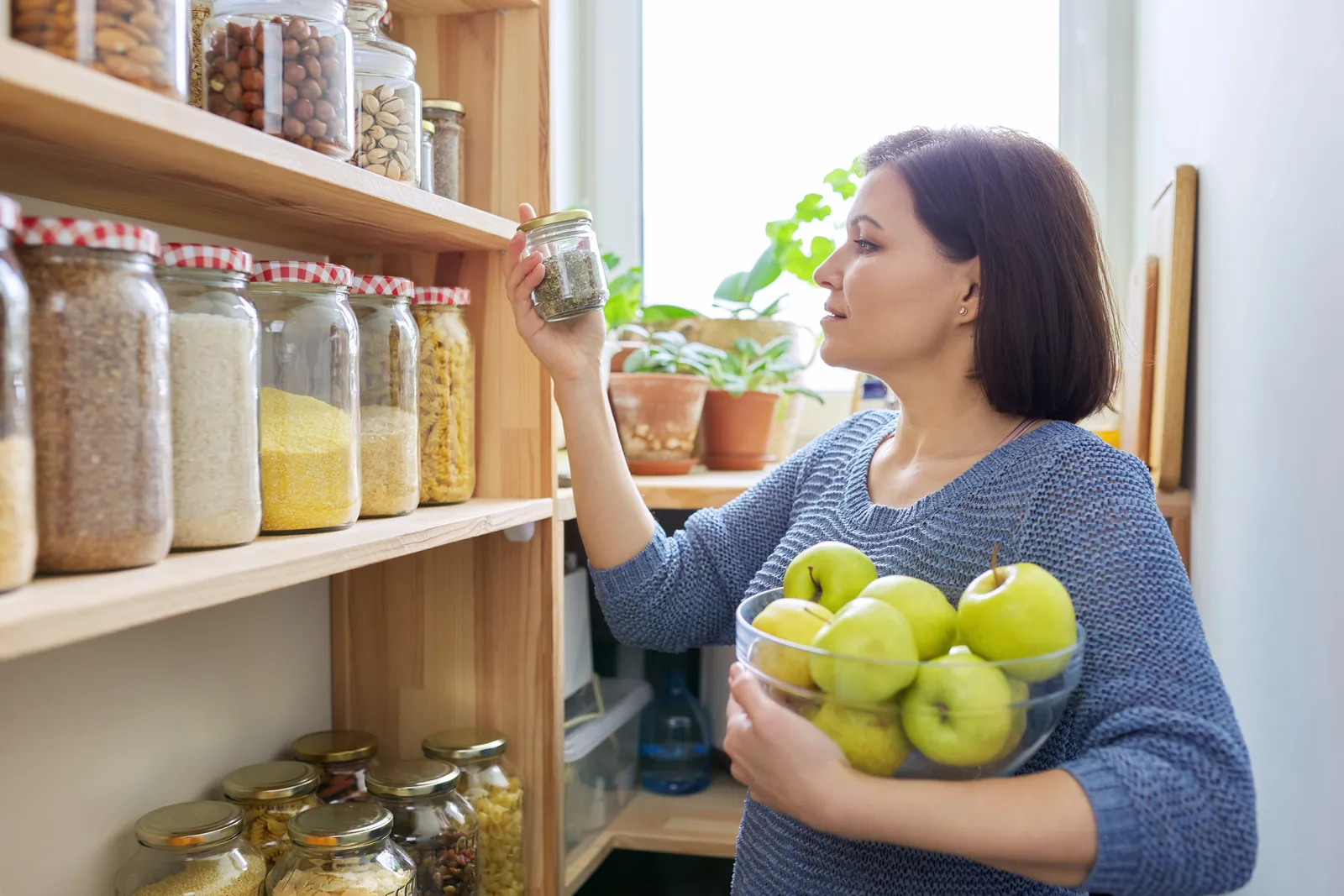 A woman holding a bowl of green apples while sorting jars on her pantry shelves